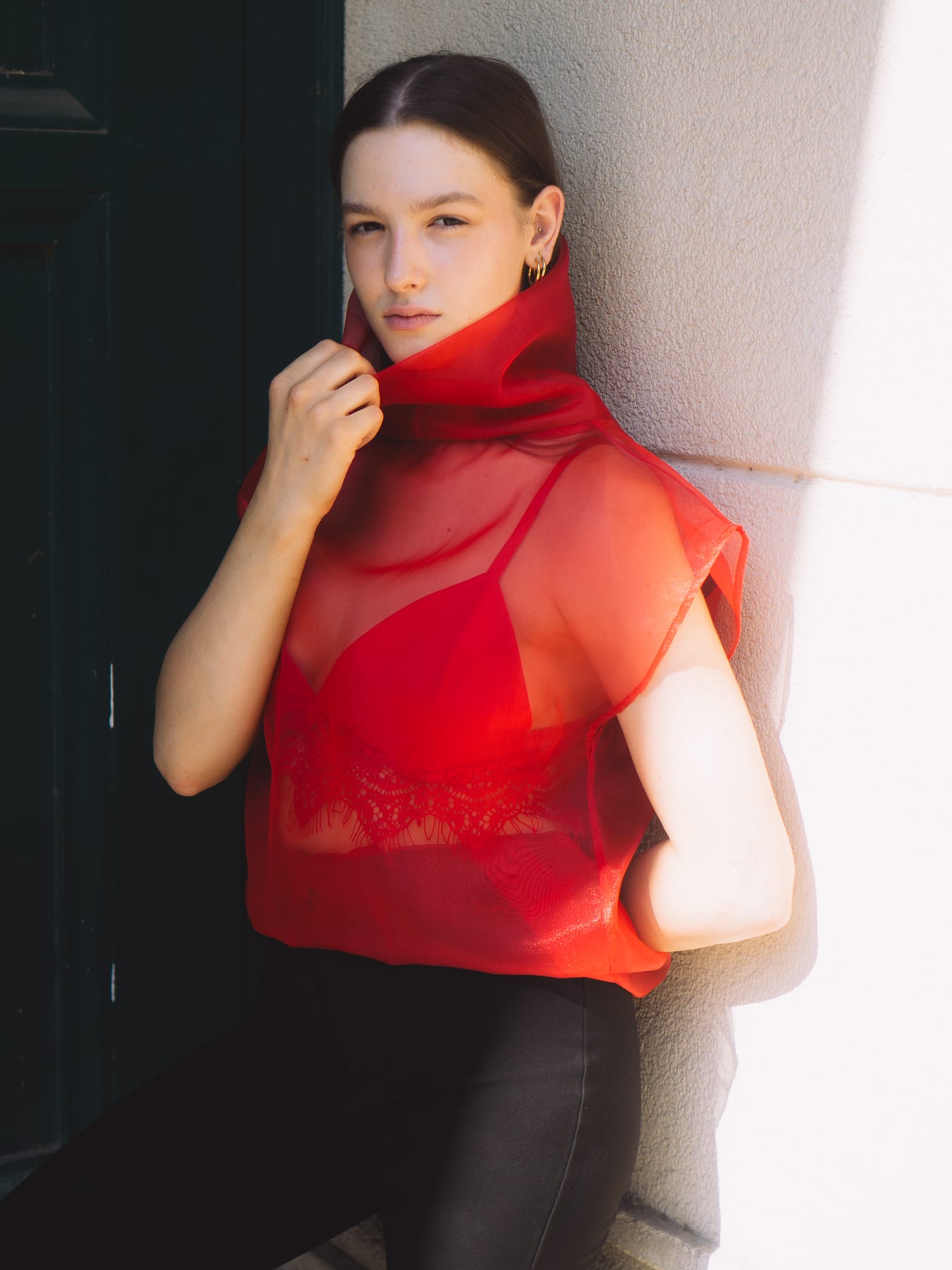 Model in a sheer red organza blouse leaning against a pale wall, holding the high collar.