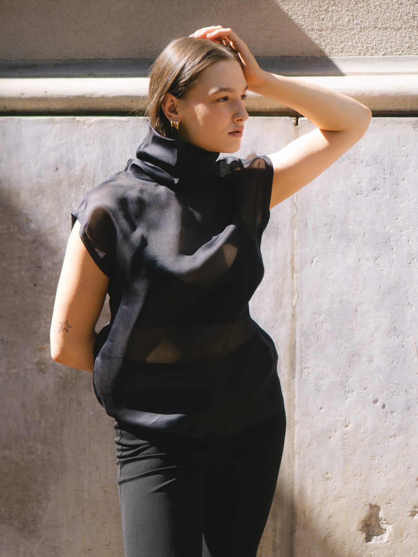 Model in a black high-neck top, one hand on her head, against a pale wall.