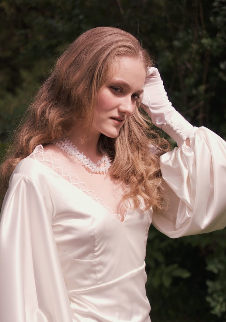 Model with long wavy blonde hair in a white satin blouse, raising a sleeve to her face.