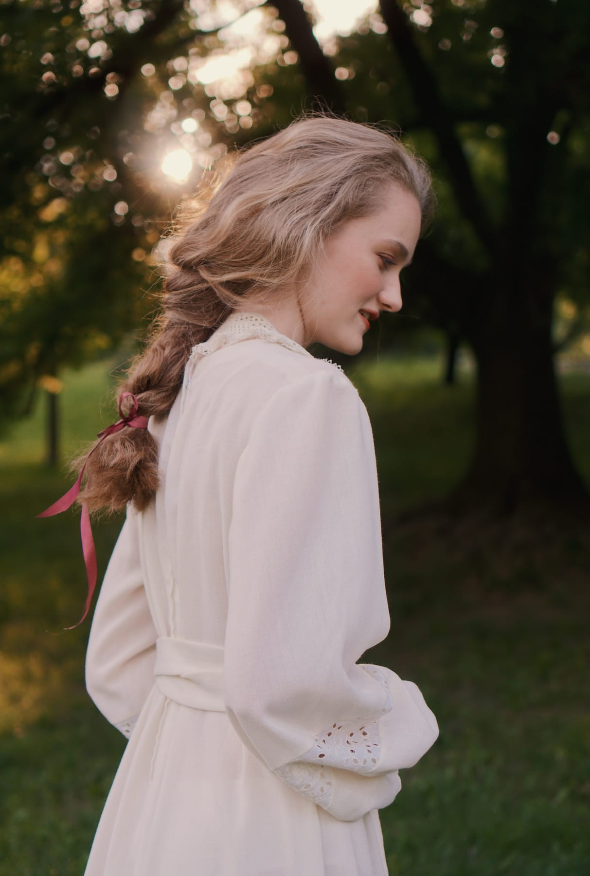 Model in a white dress among trees, seen from the side in evening light.