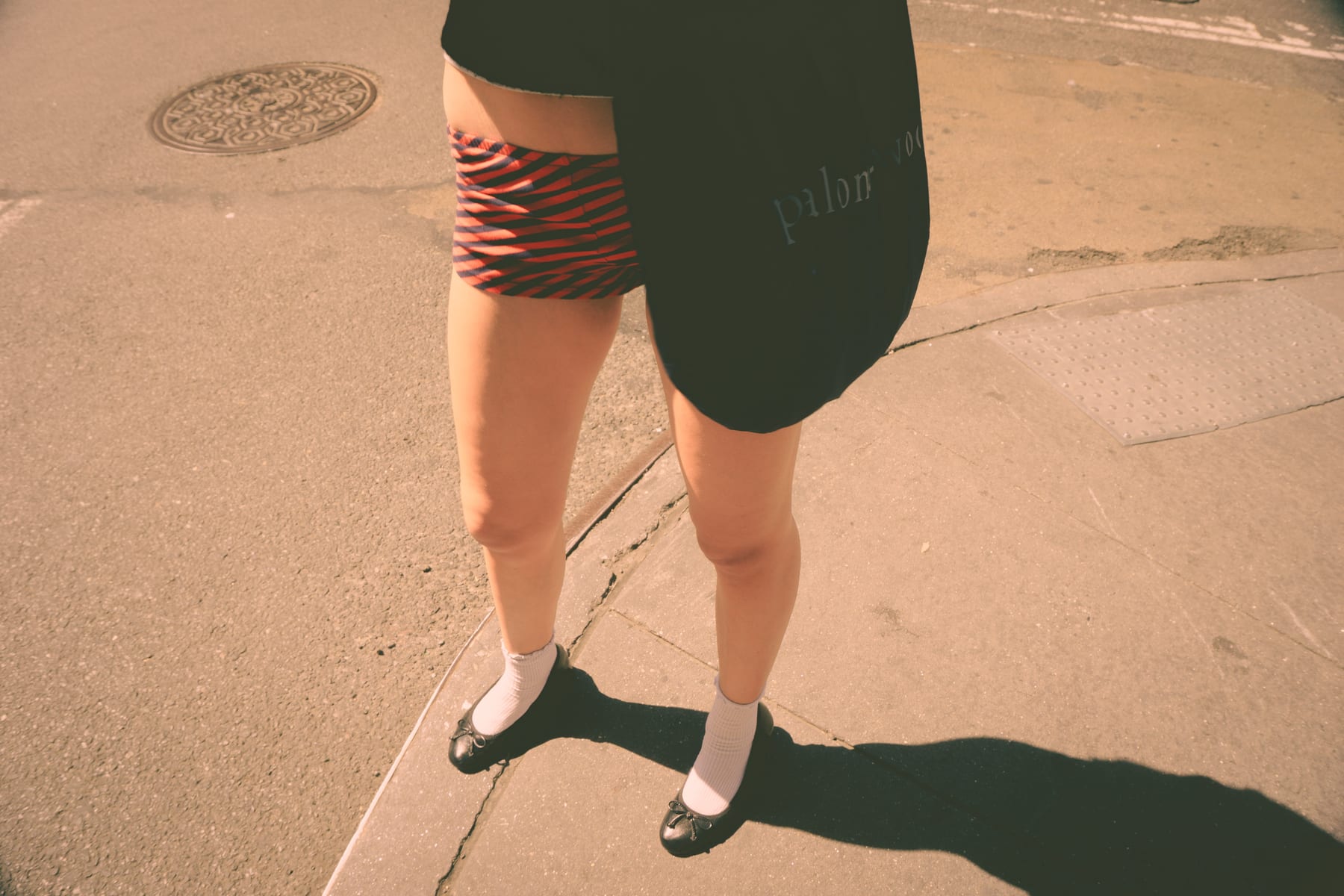 A model's legs in white socks and black shoes on a sunlit pavement, with a striped skirt and a long shadow. (dummycopy)