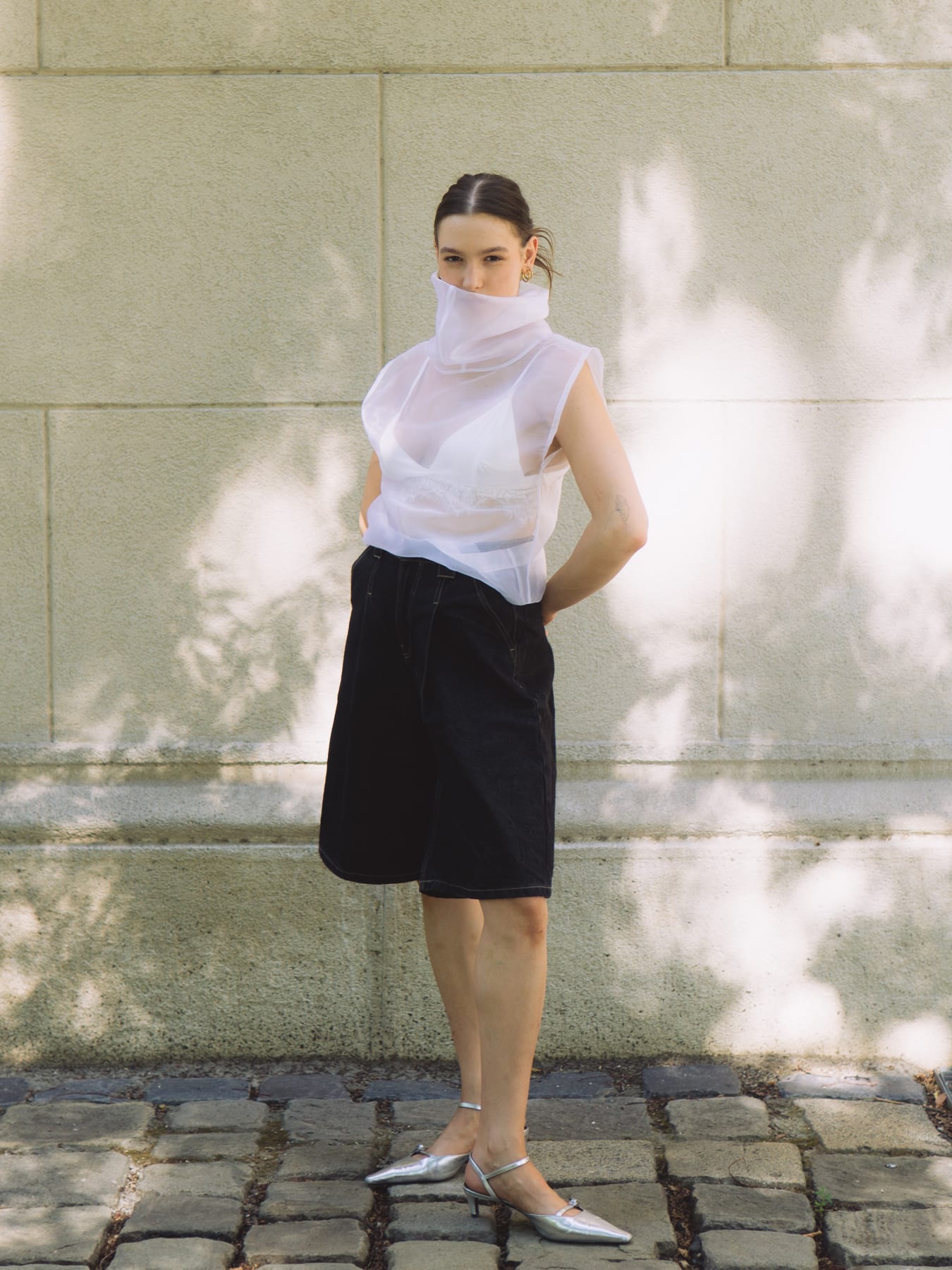 Model in a sheer white high-neck top and black denim shorts standing against a sunlit stone wall. (dummycopy)
